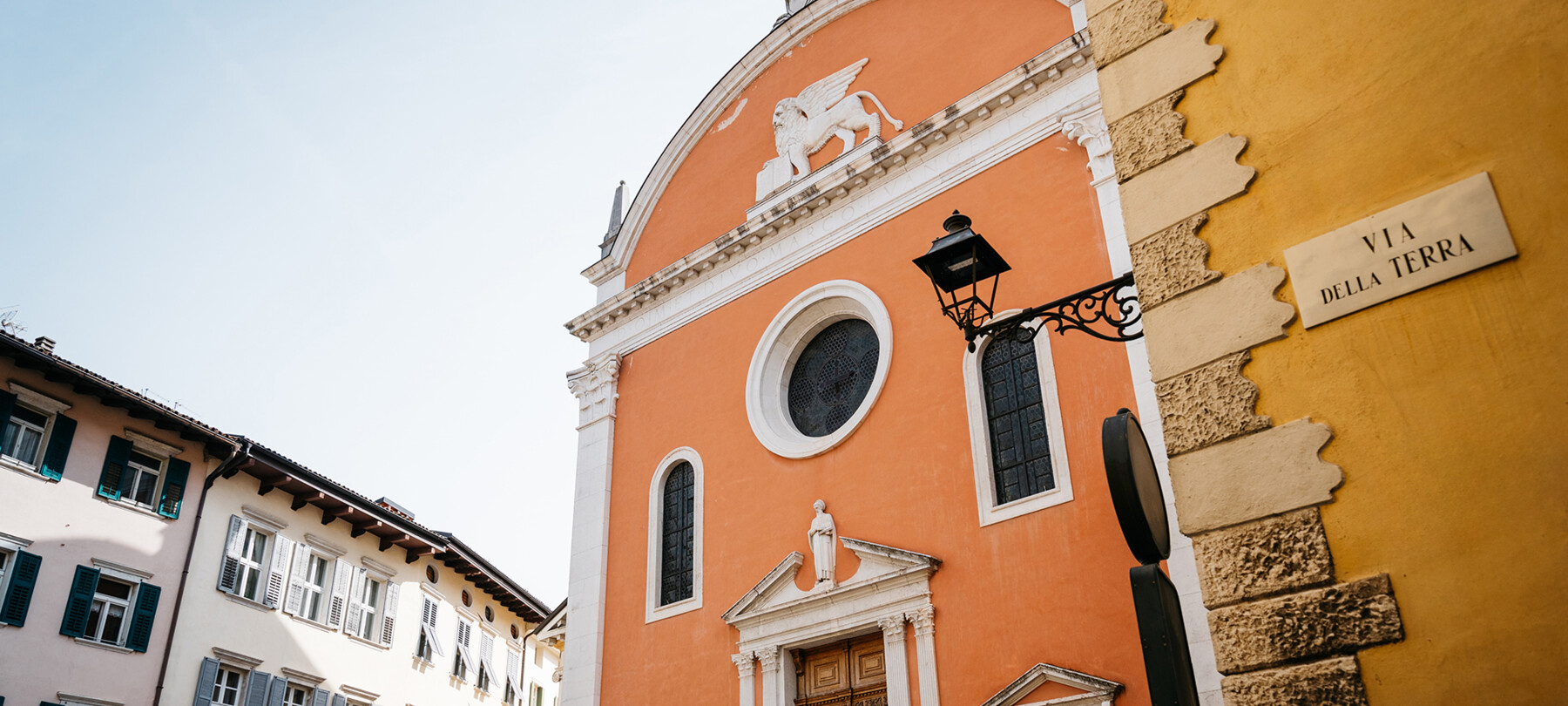 Fassade der Kirche von San Marco im historischen Zentrum von Rovereto. Die Protagonisten des Bildes sind die Farben: das Orange der Kirche, das Ockergelb des Gebäudes im Vordergrund, auf dem „Via della Terra“ steht, das Weiß und zarte Rosa der Gebäude im Hintergrund. Obwohl es nicht von Menschen bewohnt ist, ist es ein lebendiges, fröhliches Bild.
