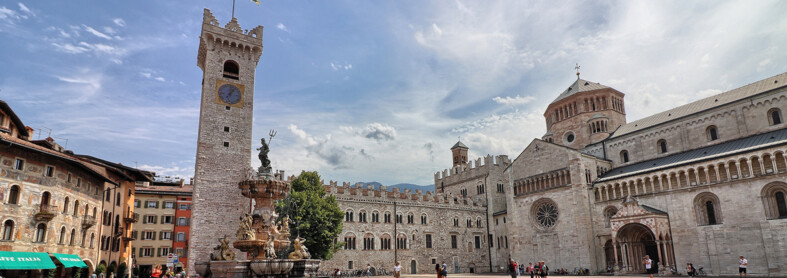 Piazza Duomo in Trento. In the background, the Civic Tower, Palazzo Pretorio and San Vigilio Cathedral, the city's cathedral. In the centre, the Neptune fountain. Behind the fountain stands the only tree in the square: a lime tree. The square, in the image, is lived in. There are some people sitting on the steps of the fountain, someone is taking photos and a small group is posing in front of the cathedral to have their picture taken. Everyone is wearing summer clothes. The blue sky is streaked with the white of light clouds.
