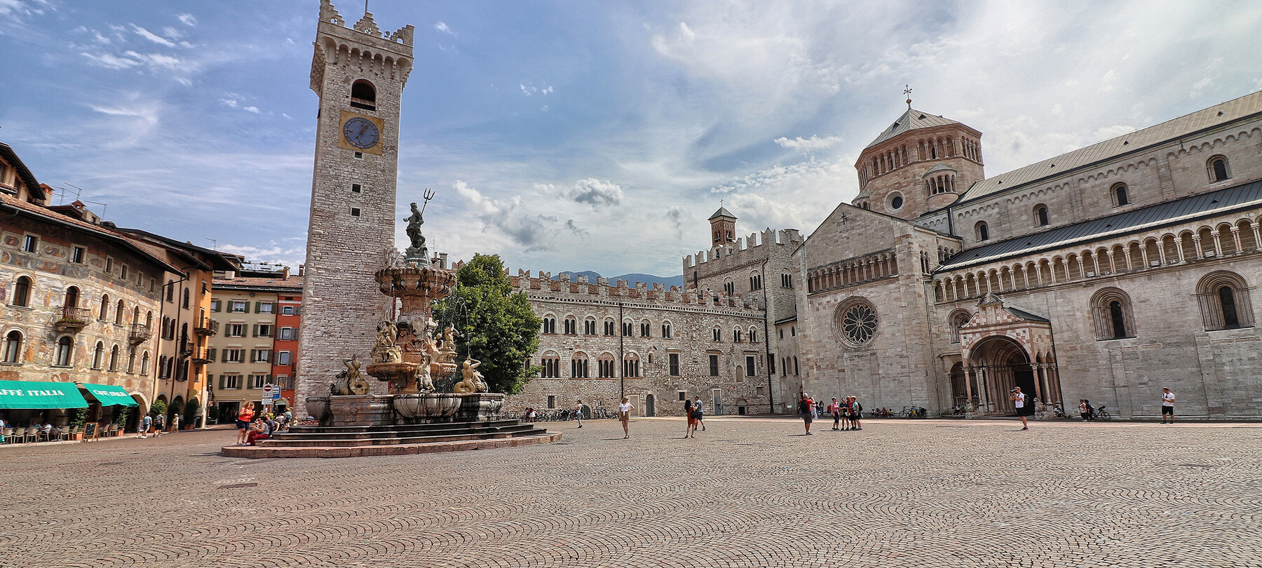 Piazza Duomo in Trient. Im Hintergrund der Stadtturm, der Palazzo Pretorio und die Kathedrale San Vigilio, die Kathedrale der Stadt. In der Mitte: der Neptunbrunnen. Hinter dem Brunnen steht der einzige Baum auf dem Platz: eine Linde. Der Platz ist auf dem Bild belebt. Einige Leute sitzen auf den Stufen des Brunnens, jemand macht Fotos und eine kleine Gruppe posiert vor der Kathedrale, um sich fotografieren zu lassen. Alle tragen Sommerkleidung. Der blaue Himmel ist mit dem Weiß leichter Wolken durchzogen.