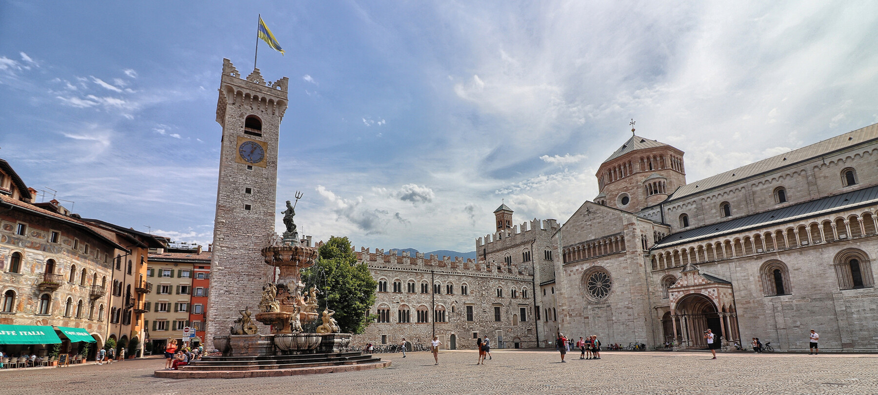 Piazza Duomo in Trento. In the background, the Civic Tower, Palazzo Pretorio and San Vigilio Cathedral, the city's cathedral. In the centre, the Neptune fountain. Behind the fountain stands the only tree in the square: a lime tree. The square, in the image, is lived in. There are some people sitting on the steps of the fountain, someone is taking photos and a small group is posing in front of the cathedral to have their picture taken. Everyone is wearing summer clothes. The blue sky is streaked with the white of light clouds.