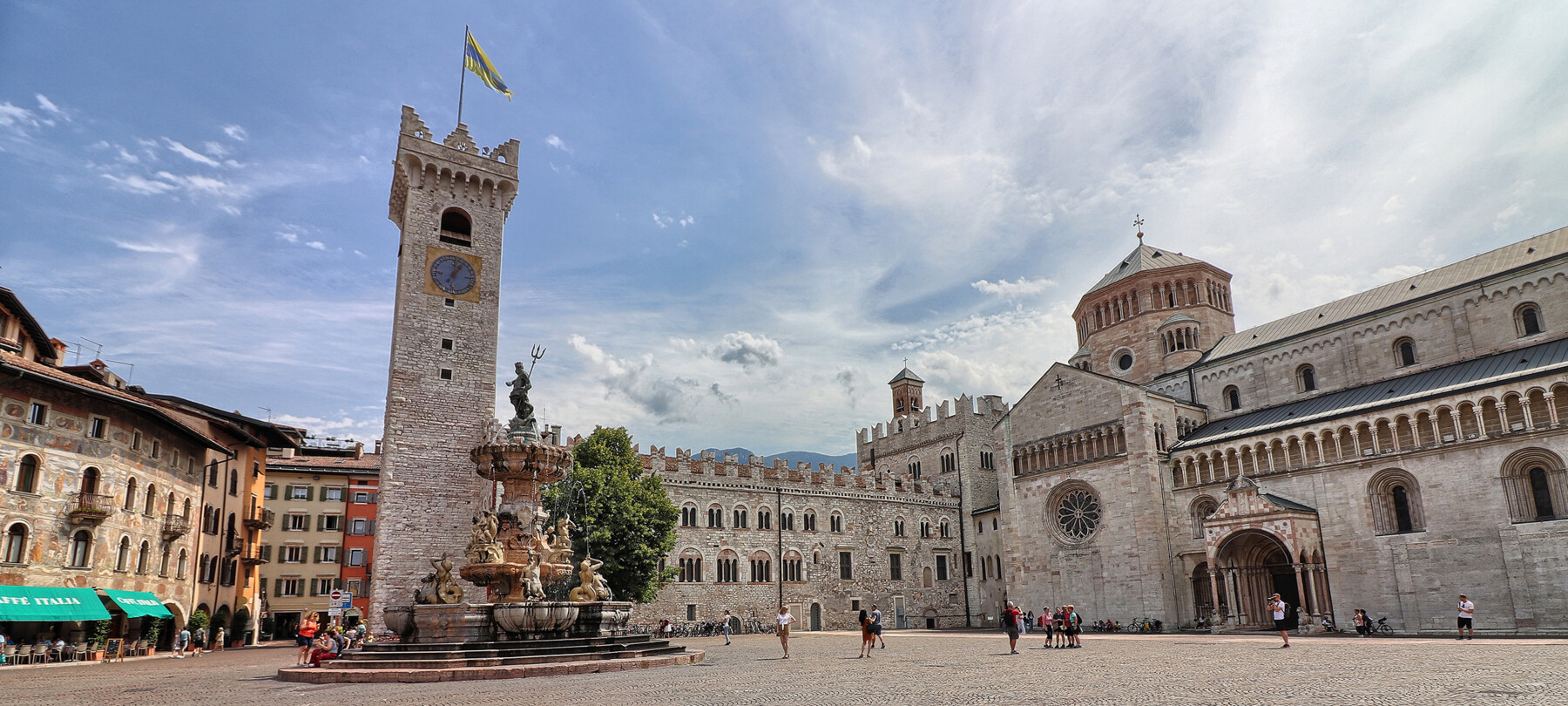 Piazza Duomo a Trento. Sullo sfondo, la Torre Civica, Palazzo Pretorio e la cattedrale di San Vigilio, Duomo della città. Al centro, la fontana del Nettuno. Dietro alla fontana svetta l’unico albero della piazza: un tiglio. La piazza, nell’immagine, è vissuta. Ci sono alcune persone sedute sui gradini della fontana, qualcuno sta scattando delle foto e un gruppetto è in posa davanti alla cattedrale per farsi fotografare. Tutti indossano abiti estivi. Il cielo azzurro è striato dal bianco di nuvole leggere.