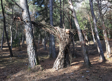 Installazione artistica di Bosco Arte Stenico. L’opera d’arte è un intreccio di rami che evoca una figura umana: le sue lunghe braccia sono distese fino ad abbracciare due alberi, uno alla sua destra e uno alla sua sinistra. In cima, un ceppo di legno più grande dei rami con cui è realizzata l’opera sembra la testa di quella figura metà uomo e metà albero.