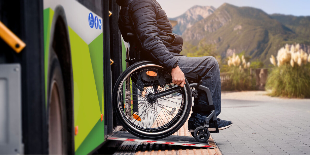 Ein Mann im Rollstuhl verlässt einen Stadtbus von Trentino Trasporti über die Rampe, die Rollstühlen den Zugang zum Fahrzeug ermöglicht. Im Hintergrund sind die Berge zu sehen, die von einem warmen, weichen Licht beleuchtet werden.