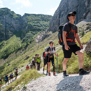 A group of people, in a line, is climbing up a trail the Brenta Dolomites. A woman, second in line, with a prosthetic left leg walks with two trekking poles, a support also used by others in the group.
