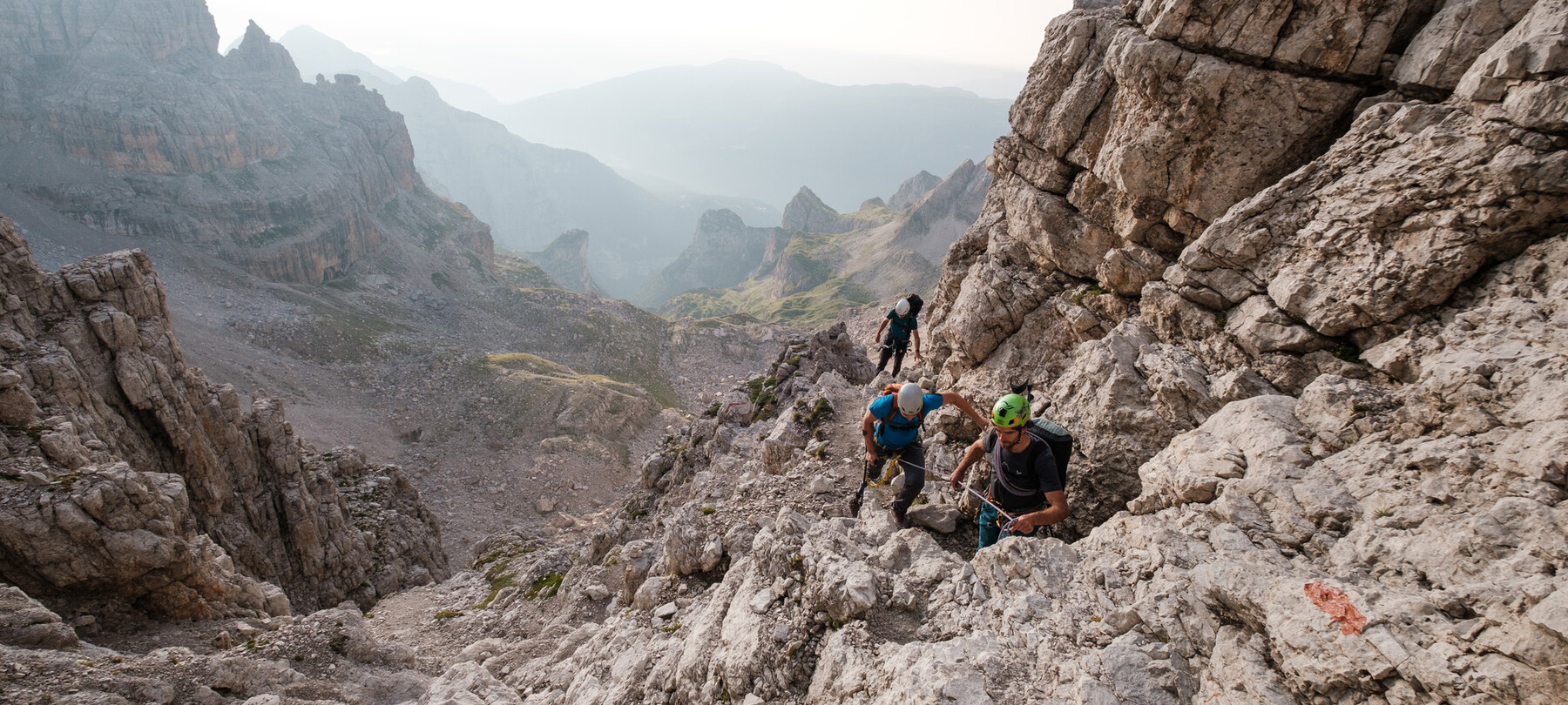 Dolomiti di Brenta, scalata verso la cima. Nel paesaggio roccioso illuminato da una luce bianca, tre alpinisti procedono in fila indiana. Uno dei tre ha una protesi alla gamba destra e scala aiutandosi con una stampella, mentre quello che apre la fila lo tiene in sicurezza con una corda che li unisce.