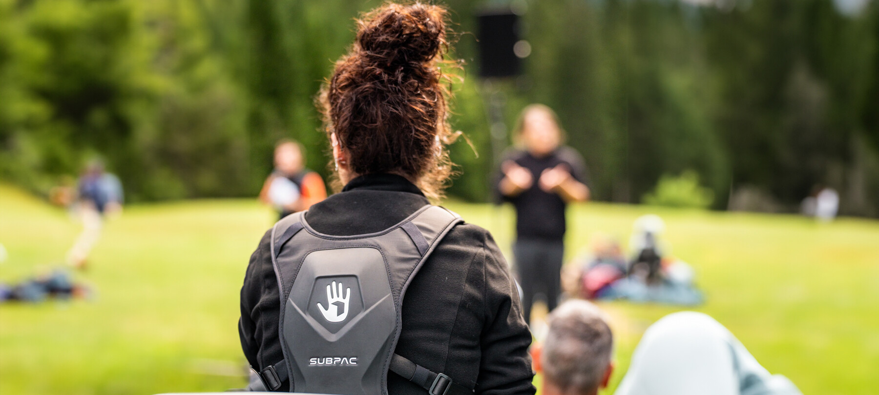 In primo piano, di spalle, una ragazza indossa uno zaino subpac. Davanti a lei, sfocati nell’immagine, gli artisti si esibiscono creando quei suoni di cui lei sentirà le vibrazioni.