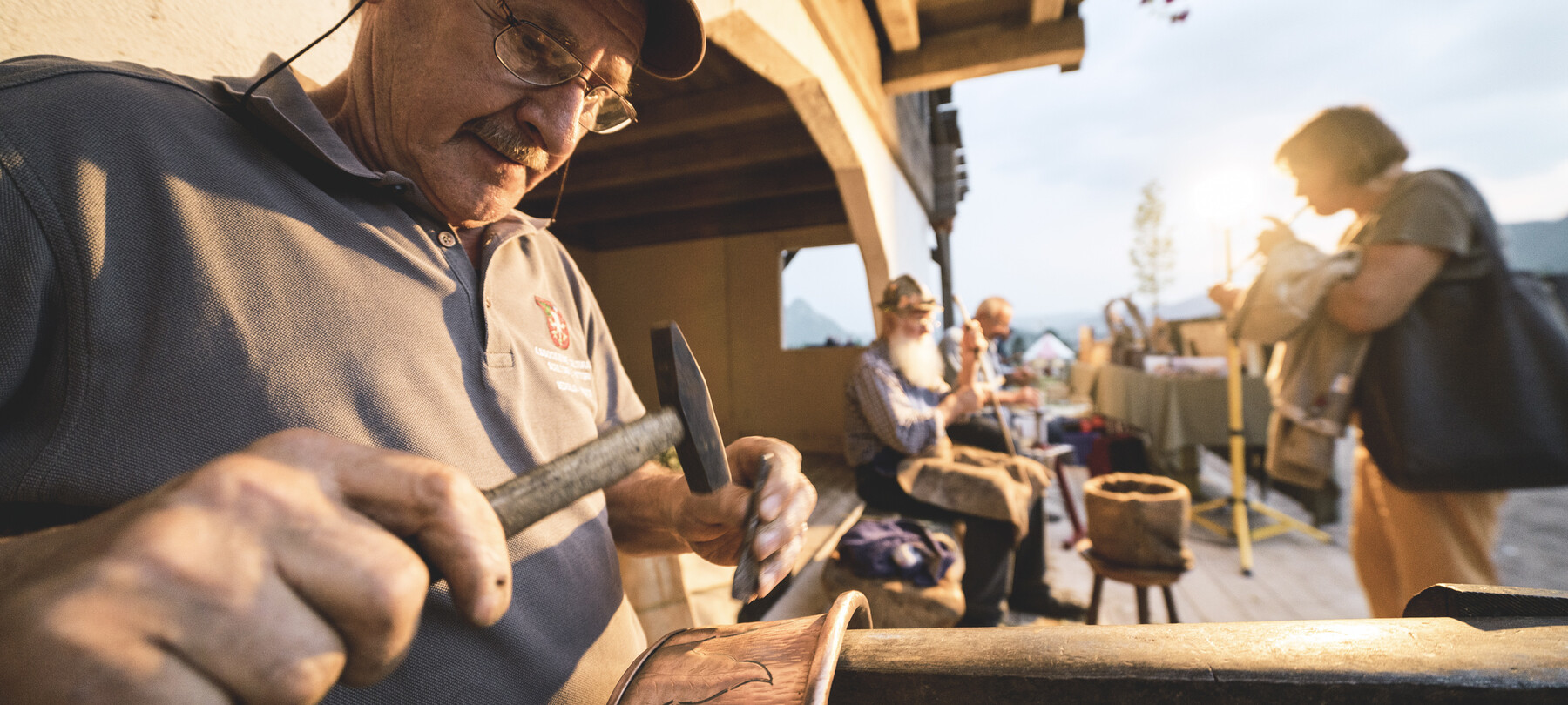 Kunsthandwerker bei der Arbeit während der Veranstaltung Tutti #fuori (alle #nachdraußen), einem Wanderfestival der Pro Loco im Trentino. Im Vordergrund graviert ein Mann Verzierungen in einen Kupfertopf. Die warme Abendsonne taucht Dinge und Menschen in goldene Reflexe.