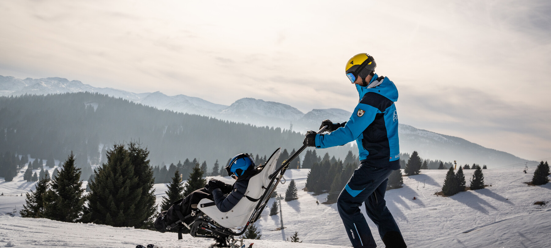 Sci inclusivo in Alpe Cimbra. Una persona con disabilità motoria scia grazie al bi-sci e al supporto di un istruttore specializzato. La luce del sole è morbida e le ombre sulla neve iniziano ad allungarsi: il tramonto non dev’essere lontano.  Nel video, persone con diverse disabilità vivono l’Alpe Cimbra sperimentando varie attività outdoor. Appassionati di biking percorrono ciclabili sbarrierate, sentieri erbosi e attraversano torrenti grazie a handbike adatte alle avventure fuori strada. Chi ama gli sport invernali, invece, si dedica alle attività sulla neve grazie ad attrezzature adatte alle necessità delle persone disabili e agli istruttori specializzati che li accompagnano nelle discese. 