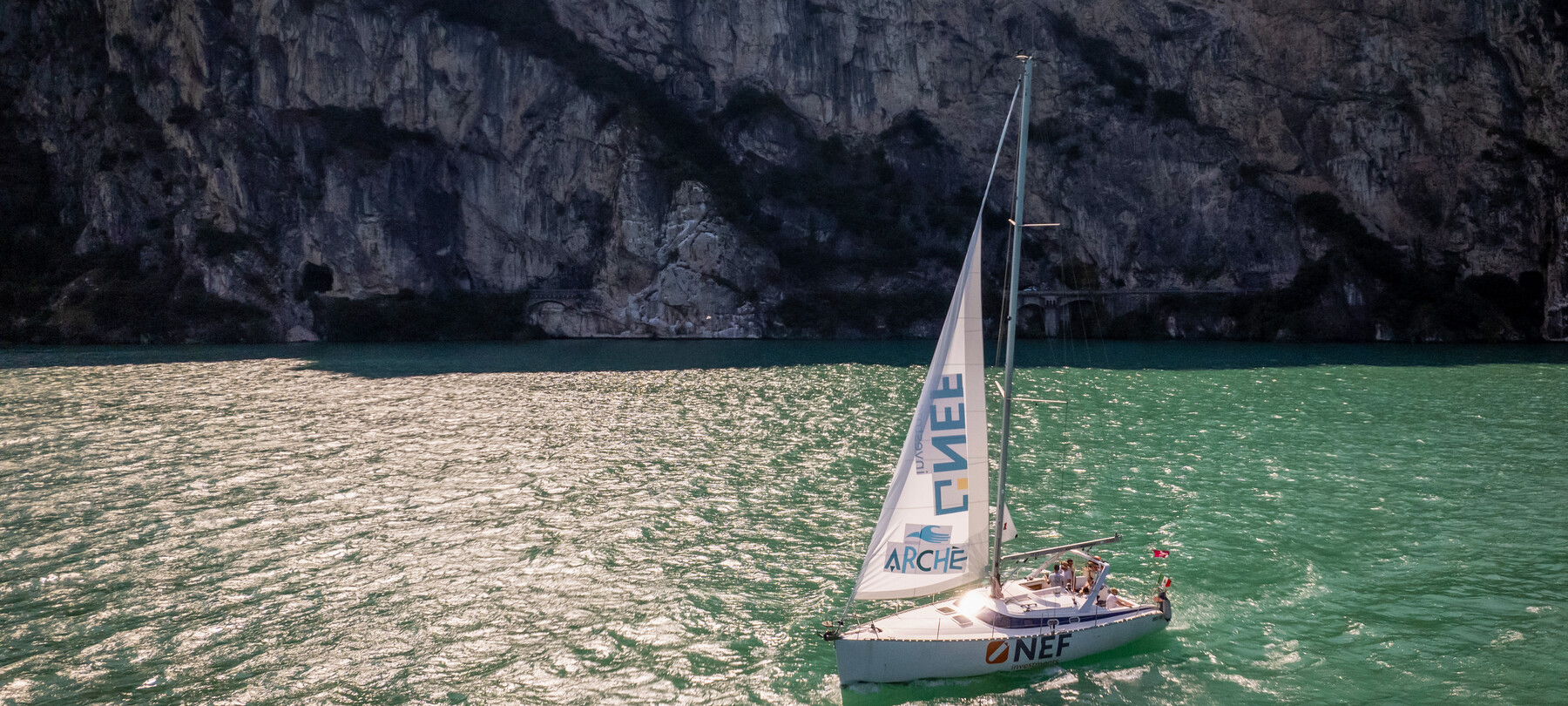 Una barca a vela della Cooperativa Archè attraversa le acque verdi e illuminate dal sole del lago di Garda. Alle spalle, una parete rocciosa in ombra chiude la scena come un sipario. | © ©Trentino Marketing_Gianluca Prati_Accessibilita_Riva del Garda_2024_00001