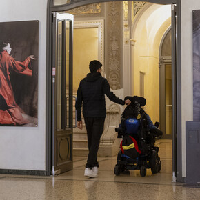 Una delle sedi del Festival dell’Economia. Due persone, di cui una in carrozzina elettrica, attraversano lo spazio, una sala dai fregi eleganti e con grandi dipinti appesi ai lati della porta che i ragazzi stanno oltrepassando.  | © ©Trentino Marketing_Origami Videography_Accessibilita_Festival Economia_2024_00092