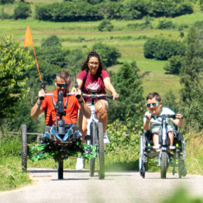 Sulla Ciclabile delle Dolomiti, tre persone si divertono facendo biking. Sono una donna, un uomo e un ragazzino: la donna cavalca una mountain-bike classica mentre l’uomo e il ragazzino guidano una handbike. Tutti e tre sorridono, l’immagine trasmette l’allegria del momento. | © ©Trentino Marketing_Gianluca Prati_Accessibilita_Val di Fiemme_2024_00026