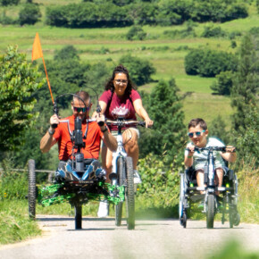 Sulla Ciclabile delle Dolomiti, tre persone si divertono facendo biking. Sono una donna, un uomo e un ragazzino: la donna cavalca una mountain-bike classica mentre l’uomo e il ragazzino guidano una handbike. Tutti e tre sorridono, l’immagine trasmette l’allegria del momento. | © ©Trentino Marketing_Gianluca Prati_Accessibilita_Val di Fiemme_2024_00026