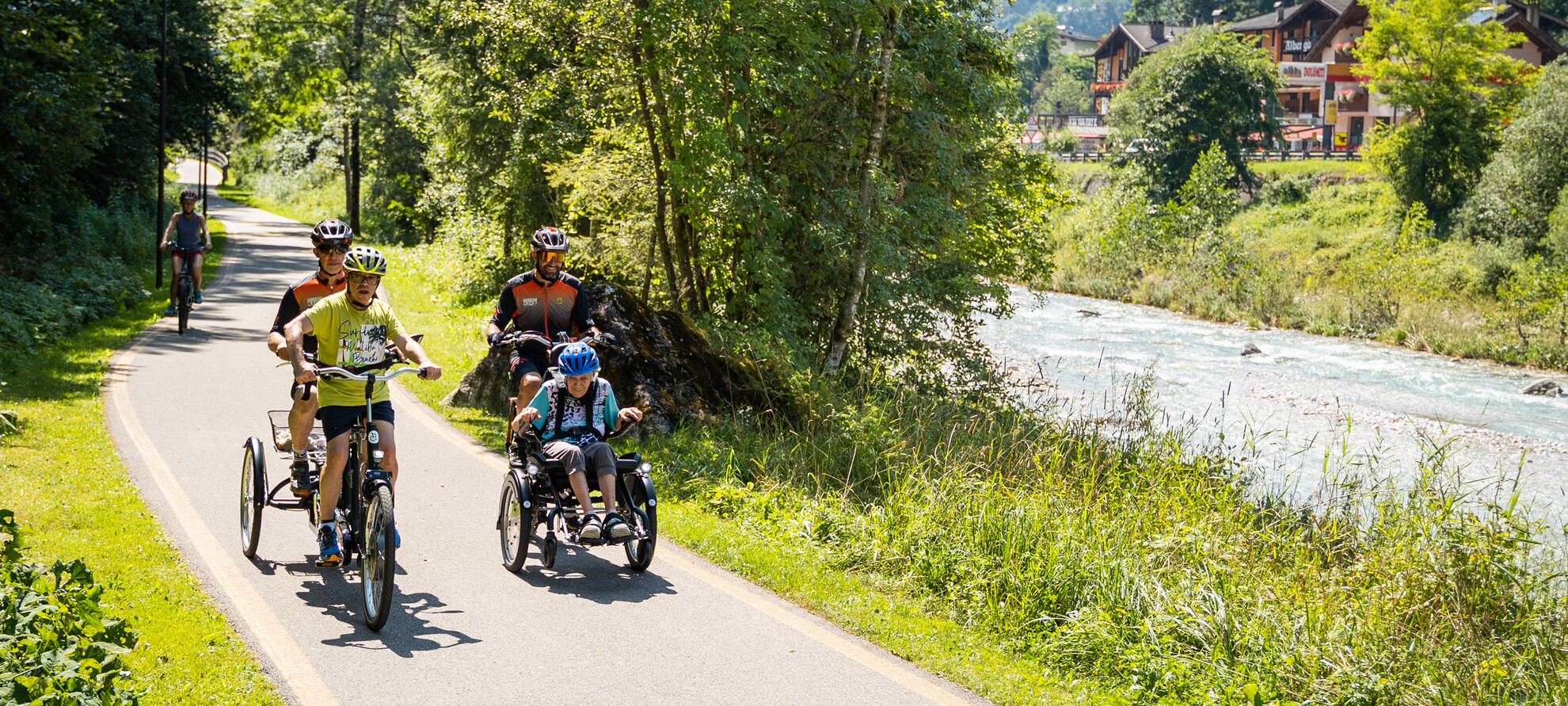 Una pista ciclabile sbarrierata costeggia un corso d’acqua. È una giornata estiva, il sole fa risplendere il verde degli alberi e si riflette sul sentiero asfaltato. Un piccolo gruppo di persone percorre la ciclabile con mezzi inclusivi: sono due tandem, di cui uno progettato per chi si muove in carrozzina. Sullo sfondo, una persona in mountain-bike segue il gruppo a distanza. | © ©Trentino Marketing_Gianluca Prati_Accessibilita_San Martino di Castrozza_2024_00036