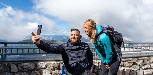 Un uomo e una donna in abbigliamento sportivo si fanno una foto dal punto panoramico del Sass Pordoi. Dietro di loro, sullo sfondo, le cime delle Dolomiti sono coperte dalle nuvole che lasciano intravedere l’azzurro del cielo.  | © ©Trentino Marketing_Gianluca Prati_Accessibilita_Val di Fassa_2024_00024