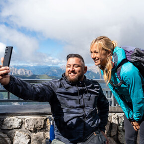 Un uomo e una donna in abbigliamento sportivo si fanno una foto dal punto panoramico del Sass Pordoi. Dietro di loro, sullo sfondo, le cime delle Dolomiti sono coperte dalle nuvole che lasciano intravedere l’azzurro del cielo.  | © ©Trentino Marketing_Gianluca Prati_Accessibilita_Val di Fassa_2024_00024