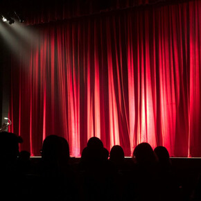 Palco teatrale con sipario rosso chiuso e luci di scena, pubblico seduto in platea.