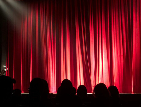 Palco teatrale con sipario rosso chiuso e luci di scena, pubblico seduto in platea.