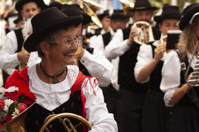 The photograph portrays a group of musicians in traditional costume, likely during a folk festival. In the foreground, an elderly woman smiles, wearing a red and white dress with a black hat, holding a golden horn and a bouquet of red and white flowers. In the background, other musicians in similar attire play instruments and wear dark hats. The atmosphere is festive and cheerful, with lights and decorations suggesting an outdoor event in a square or street. The image conveys a strong sense of tradition, community, and shared joy.