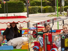 The image shows a red toy train with four children sitting on it, having fun. The train is working and moving along small tracks in the town square.