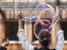 A street artist creates enormous iridescent soap bubbles, raising two large tools with his hands. In the background, a wooden structure and several people curiously observe the performance. The bubbles reflect changing colors, creating a magical and dreamy atmosphere. The event seems to take place in a public space, with adults and children captivated by the ephemeral art.