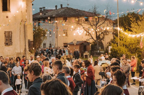 A warm and festive atmosphere fills the streets of Zambana Vecchia during Antiche Terre. The band plays surrounded by people in vintage clothing, under string lights and colorful flags. The community gathers to celebrate tradition and memory.