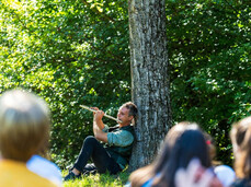 Un musicista, seduto sull'erba ai piedi di un albero in un bosco, suona un flauto traverso. Di fronte a lui, un pubblico di persone, visto di schiena, è seduto per terra e ascolta il concerto all'aperto.