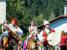 Una sfilata folkloristica anima le strade di Fai della Paganella. I partecipanti, adulti e bambini, indossano costumi tradizionali e alcuni suonano strumenti come una cornamusa e un tamburo, mentre altre persone sfilano a cavallo.