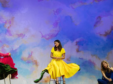On the stage, there are three women. The one in the middle is sitting on a chair and wearing a yellow dress. She is talking to the woman on the left, who is also seated on a flower-shaped chair and dressed in red. On the other side, a woman is sitting on the floor, gently touching a large artificial flower.