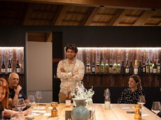 A group of people are seated at a table in the tasting room of a winery. They are enjoying themselves together with the winemaker, who is leading the tasting of his wines.