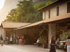 The photo shows a group of people visiting a winery in a relaxed atmosphere at sunset. Some walk through the courtyard, decorated with string lights, while others approach the entrance where the tasting is taking place. In the background, vineyards and mountains add a charming touch to the scene.