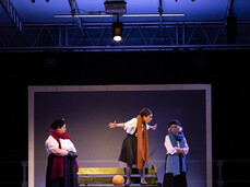 On the stage, almost completely in the dark, there are three women making faces. They are all wearing long brown skirts and colorful scarves. One of them is standing on a bench with her arms open, appearing to be angry.