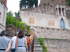 People are climbing the stairs of the hanging garden. They have headphones on their ears through which they are listening to the story of the Garden they are visiting. The terraced areas with flowers can be glimpsed, and on the right side, the citrus greenhouse is visible.