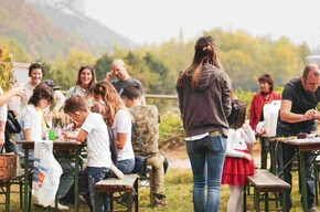 A group of children and adults take part in an outdoor activity surrounded by nature, with craft tables, games, and a warm, communal atmosphere. The event feels friendly and relaxed, ideal for a local gathering. Hills and vineyards can be seen in the background.