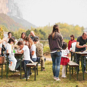 A group of children and adults take part in an outdoor activity surrounded by nature, with craft tables, games, and a warm, communal atmosphere. The event feels friendly and relaxed, ideal for a local gathering. Hills and vineyards can be seen in the background.