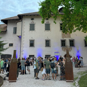 A group of people gathers in front of a historic building with walls illuminated by blue lights. The atmosphere is friendly and sociable; it is a cultural and food-and-wine event. Two metal structures bearing the word "MEZZOCORONA" mark the entrance.