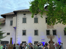 A group of people gathers in front of a historic building with walls illuminated by blue lights. The atmosphere is friendly and sociable; it is a cultural and food-and-wine event. Two metal structures bearing the word "MEZZOCORONA" mark the entrance.