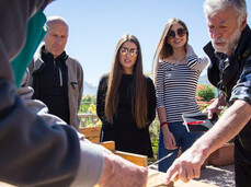 The elderly asparagus farmer is showing a group of people how to clean white asparagus. In his hand, he has a knife to remove the outer part of the asparagus. Next to him on the table are two wooden crates containing the asparagus.