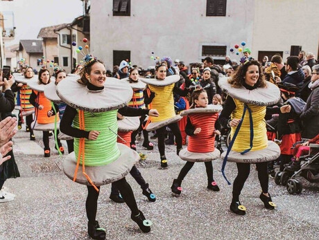 This photo captures a Carnival parade in Roveré della Luna, featuring participants dressed as colorful spools of thread. The people, including adults and children, wear creative costumes with details like threads and sewing pins. The crowd applauds and takes photos, creating a festive atmosphere. In the background, traditional village houses and other masked participants can be seen. The ground is covered in confetti, adding vibrancy to the event. The image perfectly captures the joy and energy of the celebration.