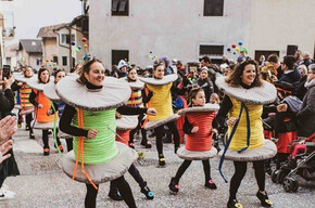 This photo captures a Carnival parade in Roveré della Luna, featuring participants dressed as colorful spools of thread. The people, including adults and children, wear creative costumes with details like threads and sewing pins. The crowd applauds and takes photos, creating a festive atmosphere. In the background, traditional village houses and other masked participants can be seen. The ground is covered in confetti, adding vibrancy to the event. The image perfectly captures the joy and energy of the celebration.