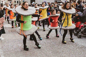 This photo captures a Carnival parade in Roveré della Luna, featuring participants dressed as colorful spools of thread. The people, including adults and children, wear creative costumes with details like threads and sewing pins. The crowd applauds and takes photos, creating a festive atmosphere. In the background, traditional village houses and other masked participants can be seen. The ground is covered in confetti, adding vibrancy to the event. The image perfectly captures the joy and energy of the celebration.