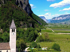 In the image, there is a small 19th-century chapel surrounded by the greenery of the surrounding vineyards. Behind it, a steep rocky cliff rises.