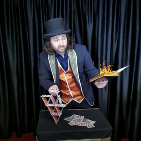 Nell'immagine si vede il mago mentre sfoglia un libro infuocato e costruisce un castello di carte