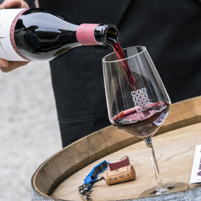 L'immagine mostra in primo piano una bottiglia di Teroldego Rotaliano che viene versato in un calice di vetro posto su una botte di legno. Molto probabilmente la bottiglia viene versata da un sommelier presente alla festa.