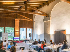 The image shows a room with exposed beams where a masterclass on Trentino wines is taking place. There are seven tables with 17 people seated, tasting multiple glasses of red wine. In the background, the speaker is discussing new grape varieties resistant to fungal diseases.