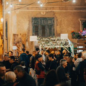 The photo shows a lively outdoor party in a historic village, illuminated by hanging lights and floral decorations. People are chatting and enjoying the convivial atmosphere, with stalls and decorated arches. The ancient walls and wooden shutters add rustic charm.