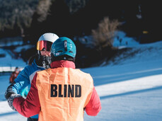 A visually impaired skier, identifiable by the orange bib with the word "BLIND" on it, is guided by a companion on a snowy ski slope.