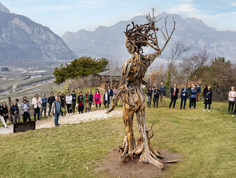 Un gruppo di persone è riunito all’aperto su un prato verde, disposto in cerchio attorno a una grande scultura in legno raffigurante una figura umana stilizzata, realizzata con rami intrecciati e radici. Sullo sfondo si estende un ampio panorama di monti e vallate, con i vigneti e le case della Piana Rotaliana visibili in lontananza. Il cielo è parzialmente nuvoloso, e la luce morbida del giorno illumina la scena, creando un’atmosfera serena e contemplativa.