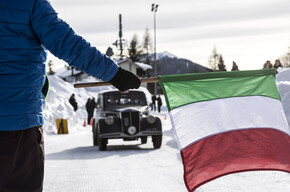 Person with Italian flag stands before a vintage car in snowy landscape.