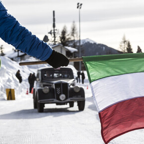 Person with Italian flag stands before a vintage car in snowy landscape.