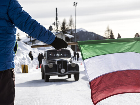 Person with Italian flag stands before a vintage car in snowy landscape.